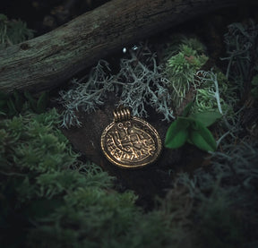 Skyddsamulett i brons med vikingrunor och brakteatdesign på snörknagg, fotograferad i naturlig skogsmiljö