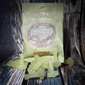 Blekgröna Mjölnir-formade godisbitar framför en pastellgul förpackning med Elder Futhark-runor och fläderblomstor. Vikinggodis i fläderblomssmaK presenterad på trä.