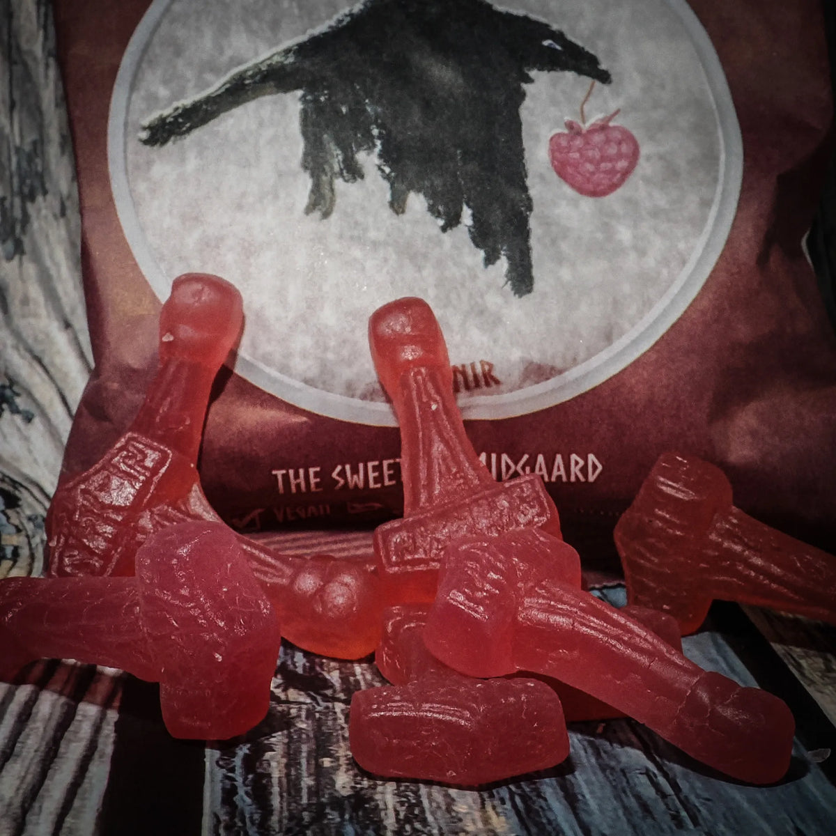 Närbild av röda hallon-smakade Mjölnirgodisar i hammarform. Flera godisar ligger på träbakgrund framför rosröd förpackning med korpillustration och texten 'The Sweets of Midgaard'.