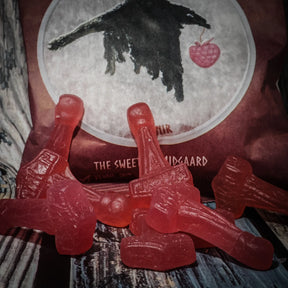 Närbild av röda hallon-smakade Mjölnirgodisar i hammarform. Flera godisar ligger på träbakgrund framför rosröd förpackning med korpillustration och texten 'The Sweets of Midgaard'.