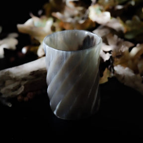 Enskilt whiskyglas i horn med spiralskuret mönster, mot mörk bakgrund med torkade löv