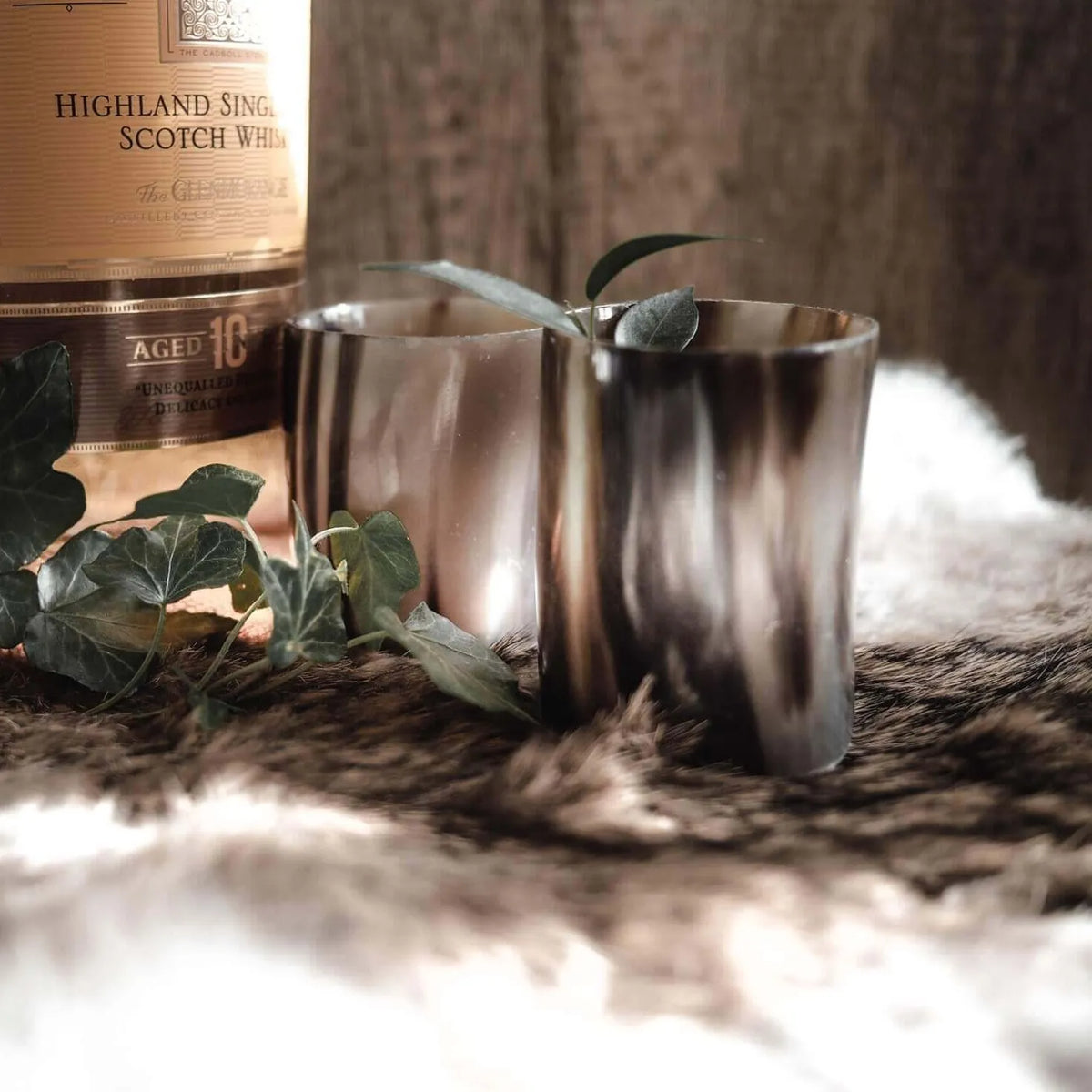 Två polerade hornglasögon för whisky bredvid en skotsk whiskybottel, dekorerad med gröna blad på naturlig bakgrund.