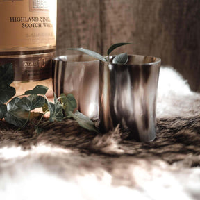 Två polerade hornglasögon för whisky bredvid en skotsk whiskybottel, dekorerad med gröna blad på naturlig bakgrund.
