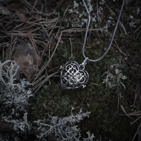 En silverfärgad änglaklocka med ornerad design, hängande från en kedja, fotograferad mot mörkare bakgrund med driftwood.