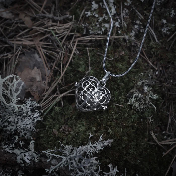 En silverfärgad änglaklocka med ornerad design, hängande från en kedja, fotograferad mot mörkare bakgrund med driftwood.