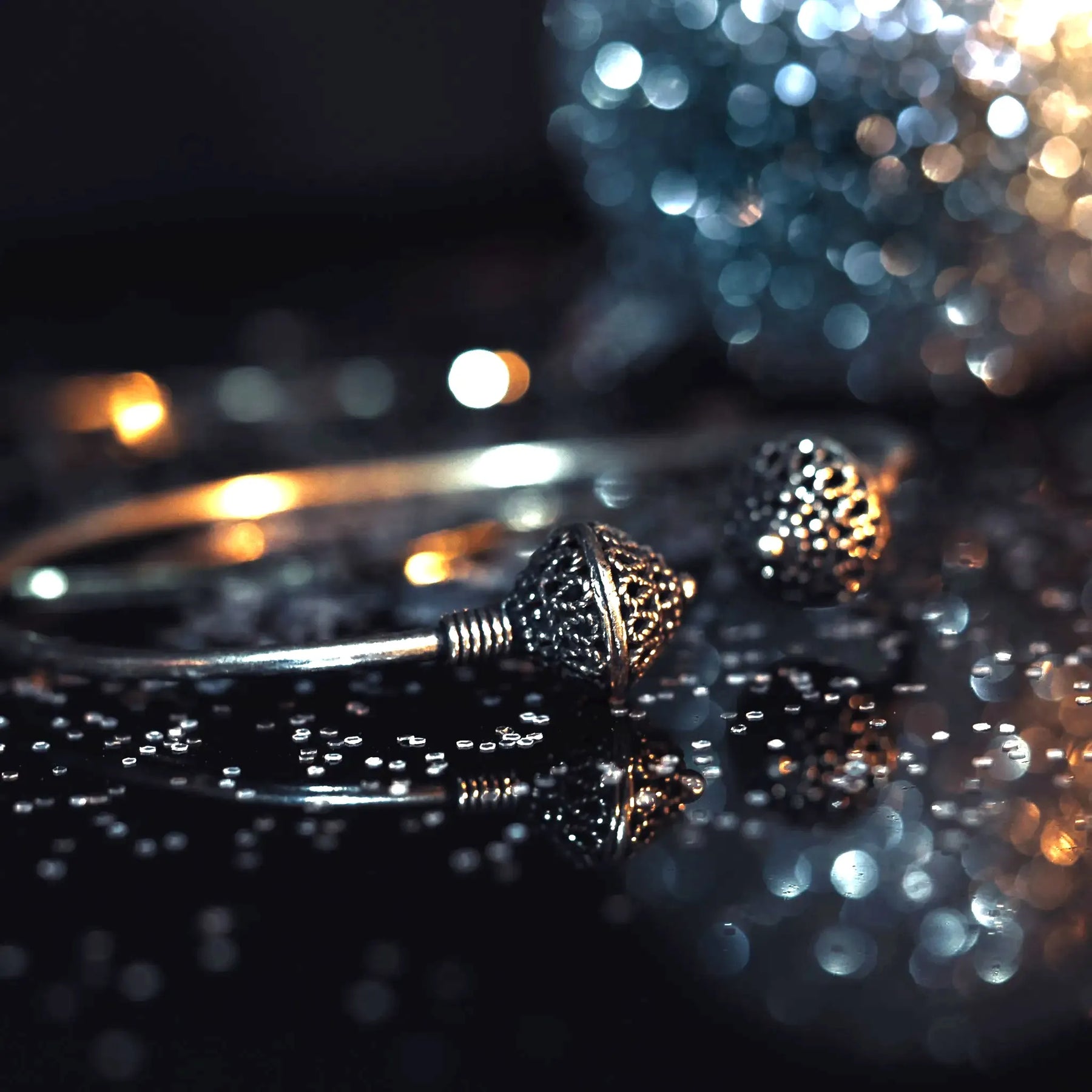 Silverpläterad mässingarmband i dramatisk studiobelysning. Två graverade pärlor med vikingatida motiv visas i närbild på reflekterande yta. Varmt och kyligt bokeh-ljus i bakgrund skapar lyxig presentationsmiljö. Tråd-lindade anslutningspunkter visar traditionell teknik.