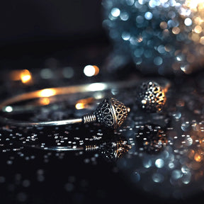 Silverpläterad mässingarmband i dramatisk studiobelysning. Två graverade pärlor med vikingatida motiv visas i närbild på reflekterande yta. Varmt och kyligt bokeh-ljus i bakgrund skapar lyxig presentationsmiljö. Tråd-lindade anslutningspunkter visar traditionell teknik.