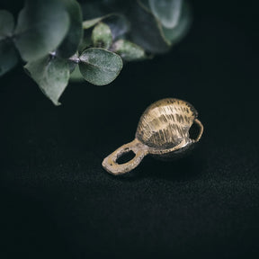 Mässingbjällra med rispig textur och elegant päronformad kropp, upphängd från en öglor mot mörk bakgrund med diskret bladverk