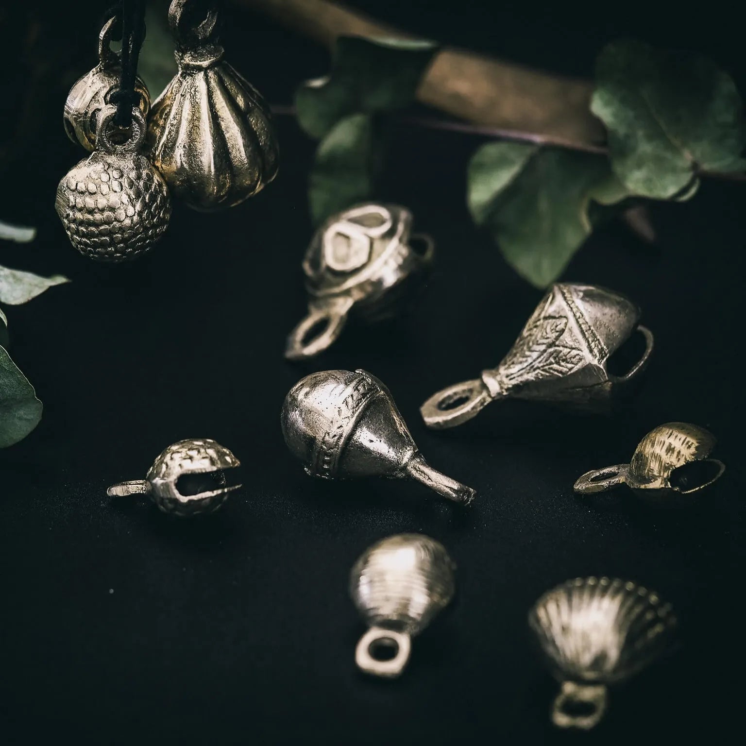 Samling av mässingbjällror i varierande storlekar med filigrébroderi, snäckornament och lövskulpterad ytterkant, arrangerade med eukalyptusblad på mörk yta