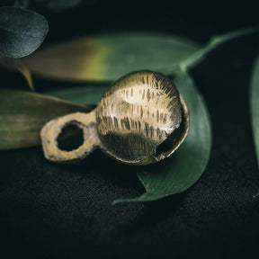 35mm mässingklocka med Rispig ytfinish. Tydliga linjära åsar bildar vågande mönster på hela klockans yta. Naturlig växtbakgrund. Studiefotografi som framhäver texturen.