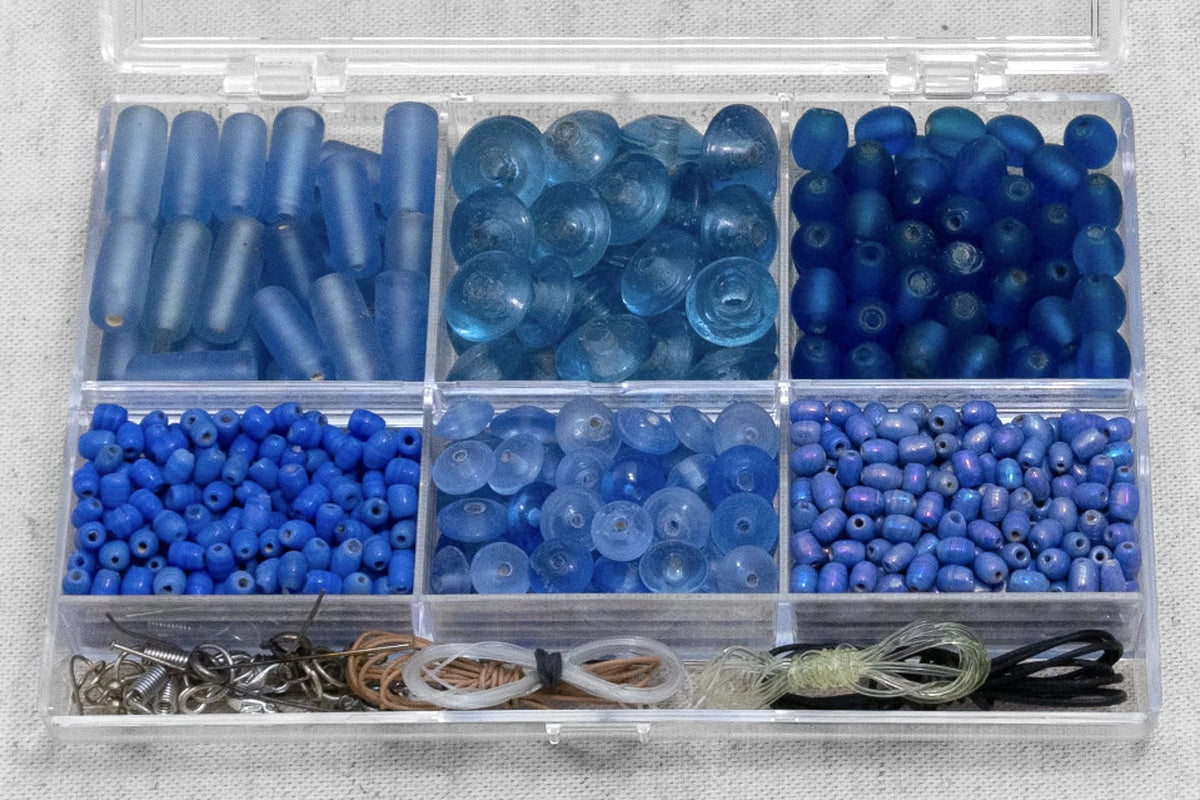 Bead box organizer containing blue glass beads in six different shades and sizes ranging from 6mm to larger faceted stones, with metal findings in bottom compartment