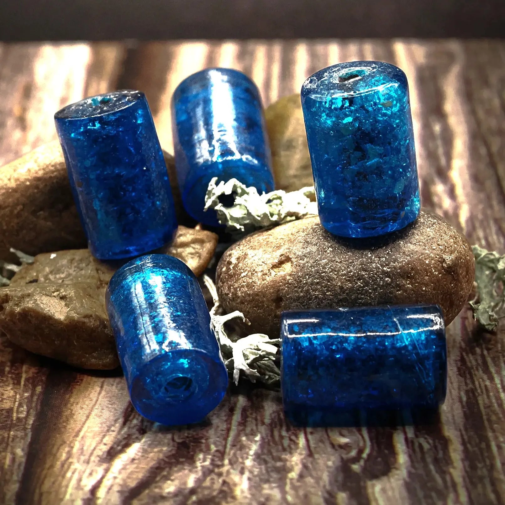 Blå cylindriska glaspärlor på stenar och träunderlag. Stora pärlor för smycken med glansig finish och metallicglans.