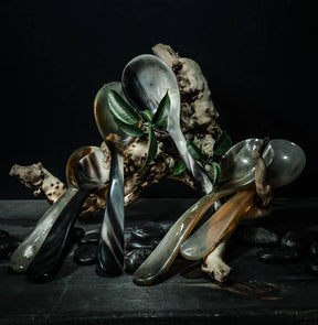 Matskedar i horn, set med 6 stycken. Varje sked visar naturlig hornstruktur med mörkgrå till ljusbeige färgtoner. Skedarna är arrangerade på en mörk trävobbyyta omgivna av naturliga träelement och gröna växter.