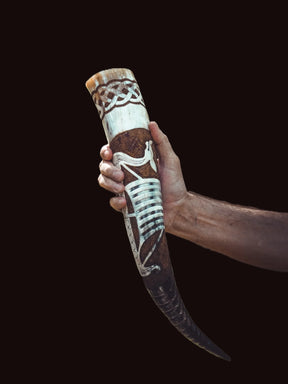 Dryckeshorn av organiskt horn hållet i hand, med graverad vikingaskepp-design och metallband. Studio-fotografi mot mörk bakgrund som visar detaljerna i handarbetet och hornets naturliga struktur.