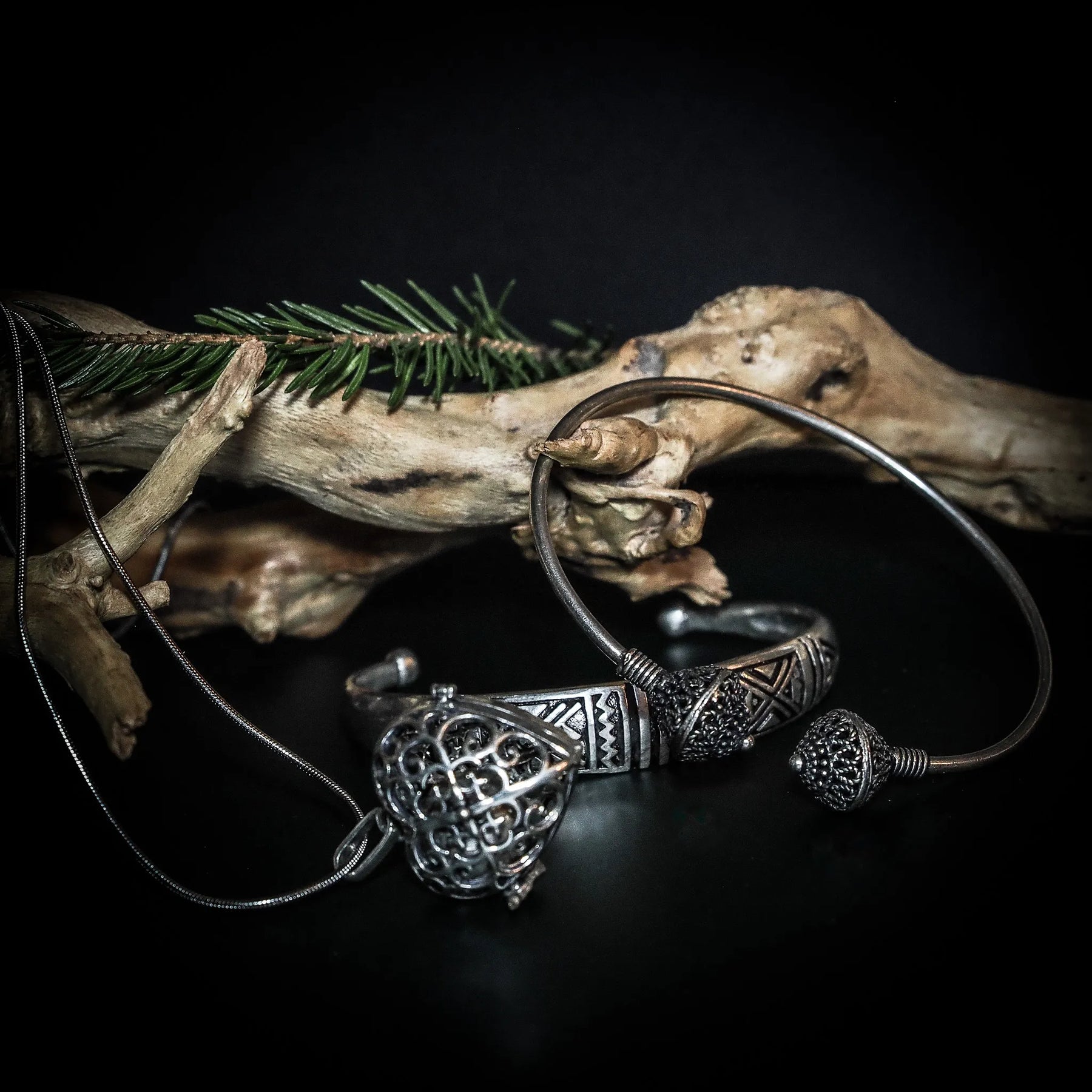 Freja gåvoset presenterat mot driftwood och grangrenar, visar silverfärgade armband med vikinginspirerade mönster, hängande knutverksmotiv och änglaklocka.
