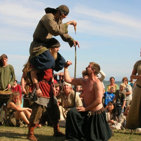 Festivalfotografi visar man lyft på axlarna av annan man i vikingakläder, omgiven av publik på gräsmatta - en klassisk vikingatid styrkelek