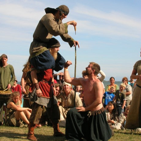 Festivalfotografi visar man lyft på axlarna av annan man i vikingakläder, omgiven av publik på gräsmatta - en klassisk vikingatid styrkelek
