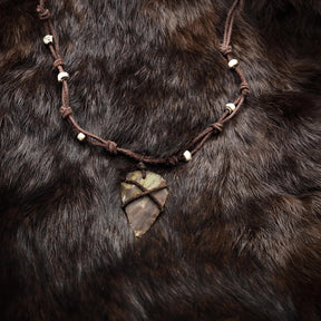 Handgjord stenpilspets i varmt ocker-ton hängande från mörkbrunt läderband dekorerat med små benärlor. Fotograferad mot mörk pälsbakgrund för att framhäva neolitisk autenticitet och vikingakänsla.