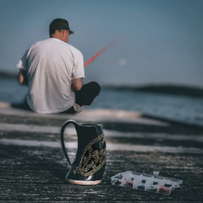 Två hornmuggar med Sigurdsagan-gravering på stranden. Naturligt hornmaterial med mörka graverade motiv. Mörkerivet hav och himmel skapar atmosfär av norsk sjöfartstradition.