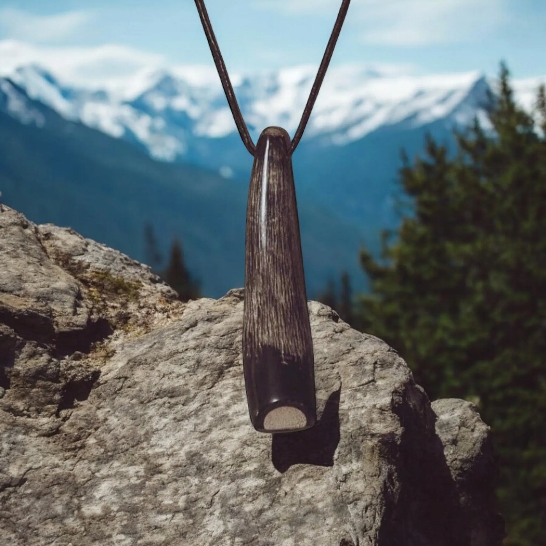Visselpipa av horn hängande på lädersnöre mot snöbetäckt alpint berglandskap med barrskog. Visar produkten i naturlig utomhusmiljö passande för vandring och nordisk överklass-aktivitet.