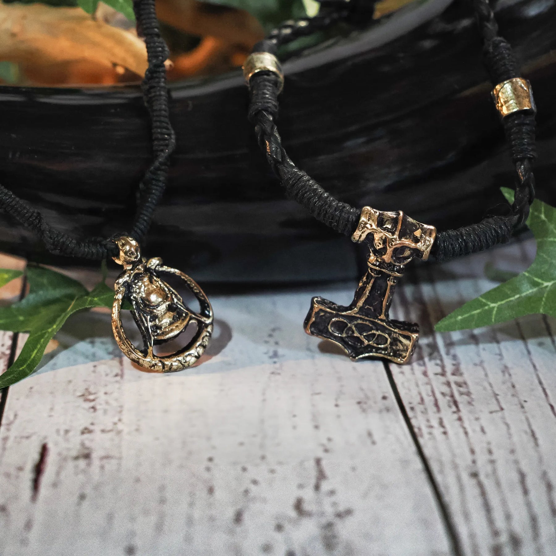 Närbild av vikingbröllopspakets smycken - svarta lädersnören med guldiga Mjölnir- och Freja-anhängen, ordnade på träyta med växtdekorationer
