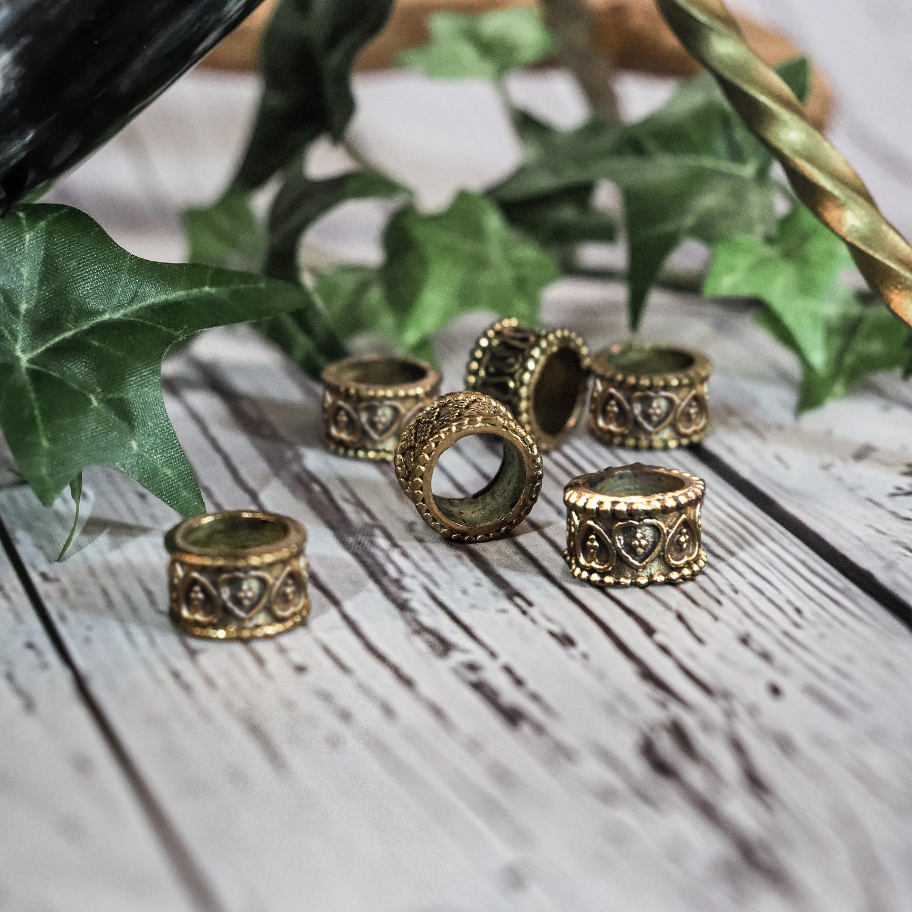 Mässingsörhängen och dekorativa pärlor med vikingainspirerad gravering, arrangerade på vit trä-bakgrund tillsammans med gröna växtdelar