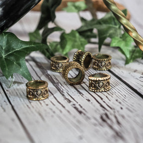 Mässingsörhängen och dekorativa pärlor med vikingainspirerad gravering, arrangerade på vit trä-bakgrund tillsammans med gröna växtdelar