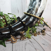 Vikingabröllops-dryckeshorn i svart läder med mässingsdetaljer, omgivet av växtdekorationer och smycken på vit trä-bakgrund