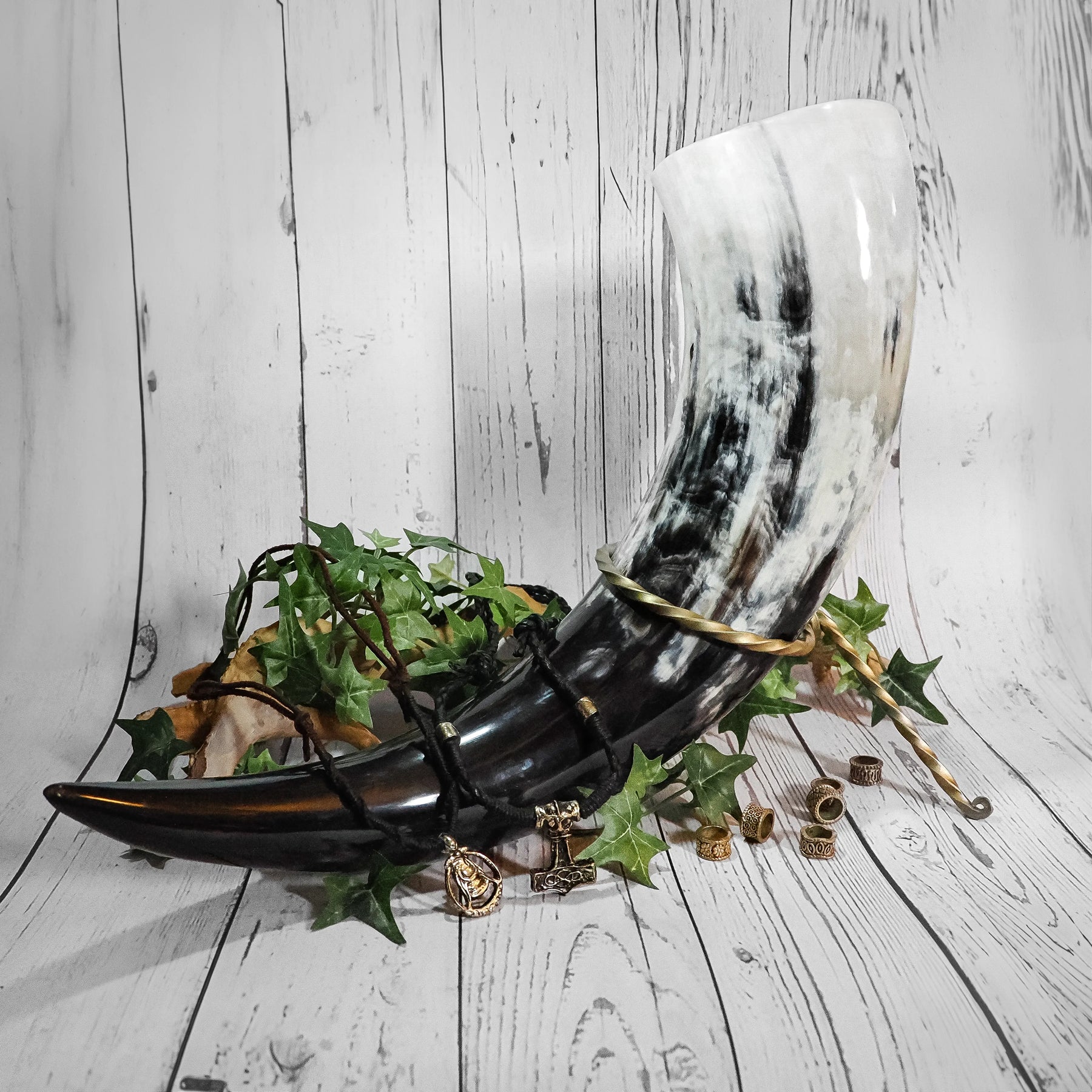 Vikingabröllops-dryckeshorn i svart läder med mässingsdetaljer, omgivet av växtdekorationer och smycken på vit trä-bakgrund
