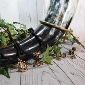 Vikingabröllops-dryckeshorn i svart läder med mässingsdetaljer, omgivet av växtdekorationer och smycken på vit trä-bakgrund