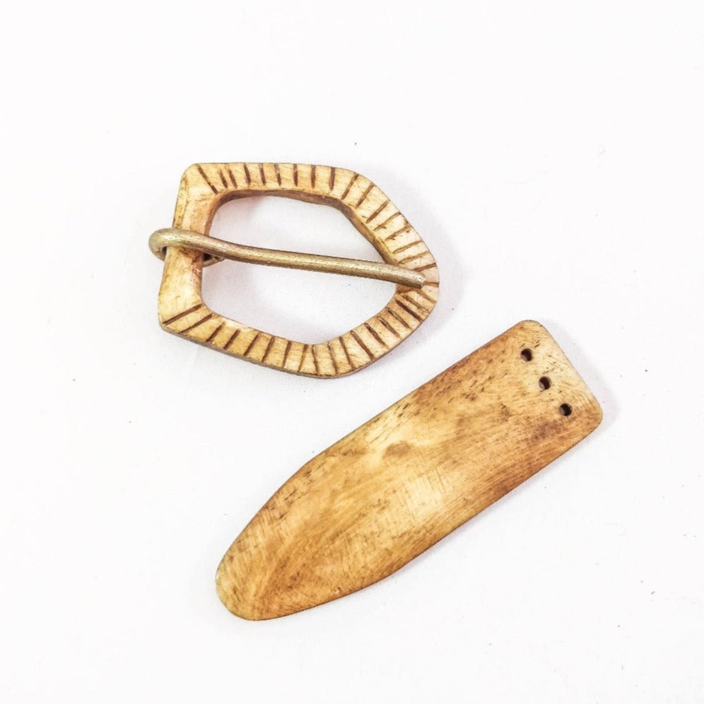 Bältesspänne i ben med två komponenter - oval spännearm med vertikalt kannelerad dekor och funktionell tungpinne, samt förlängd bältestungbit med tre fästhål på vit bakgrund