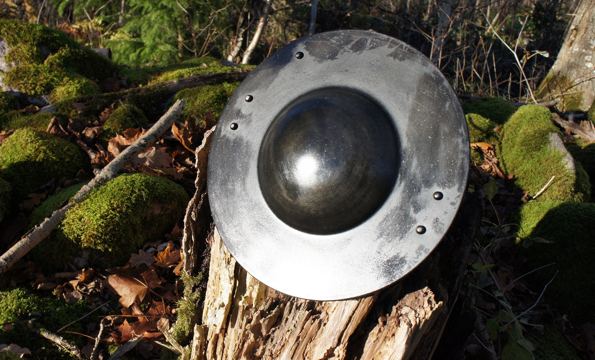 Metallbuckler sköld fotograferad framifrån, visar konvex bål och silvergrå fläck med rostig patina på naturlig skogsmark