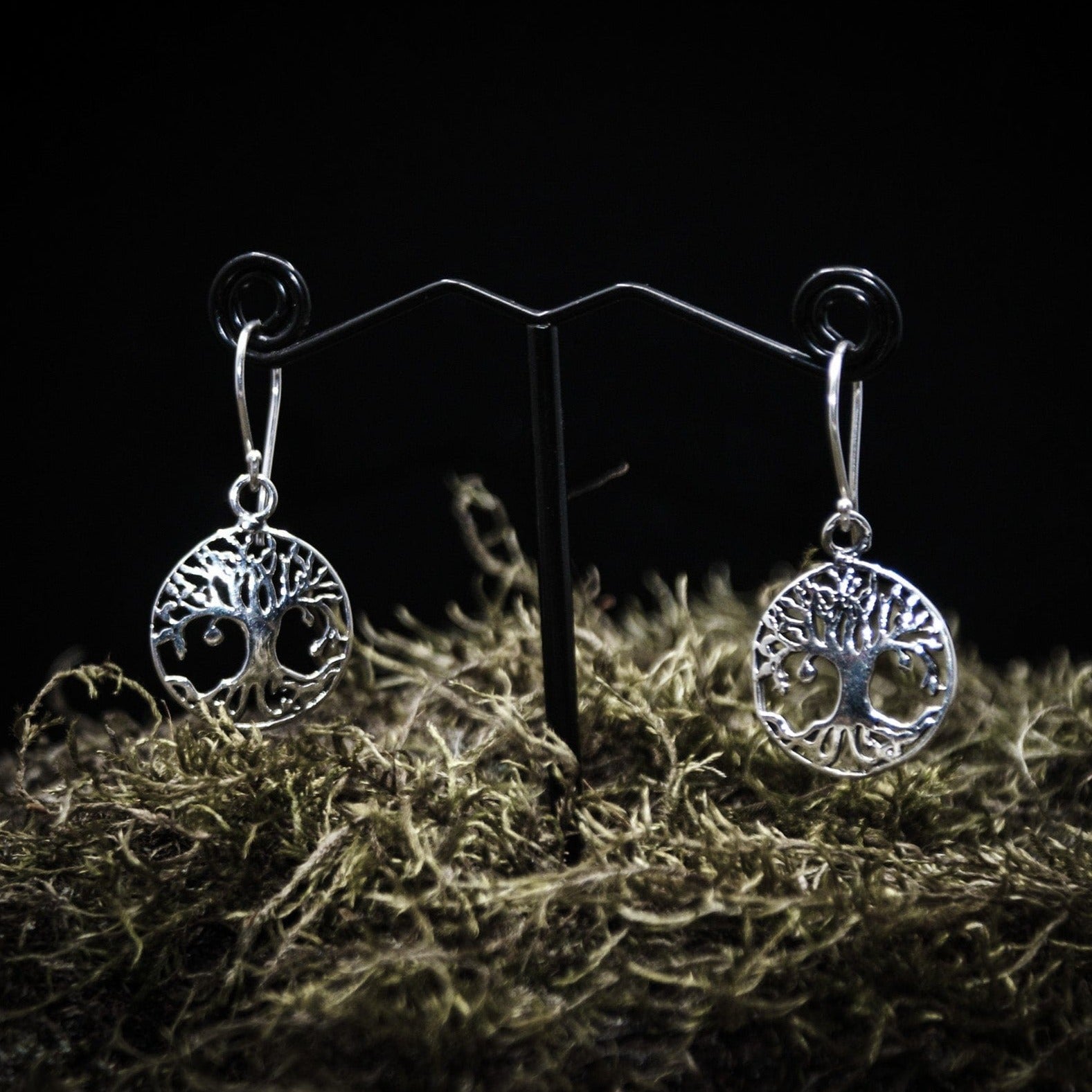 Ett par silverpläterade Yggdrasil örhängen med detaljerad träd motiv i cirkulär ram. Visade på T-ställ med mörk bakgrund och torkad växtmaterial som bas.