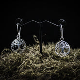 Ett par silverpläterade Yggdrasil örhängen med detaljerad träd motiv i cirkulär ram. Visade på T-ställ med mörk bakgrund och torkad växtmaterial som bas.