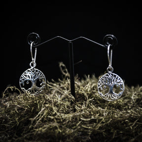 Ett par silverpläterade Yggdrasil örhängen med detaljerad träd motiv i cirkulär ram. Visade på T-ställ med mörk bakgrund och torkad växtmaterial som bas.