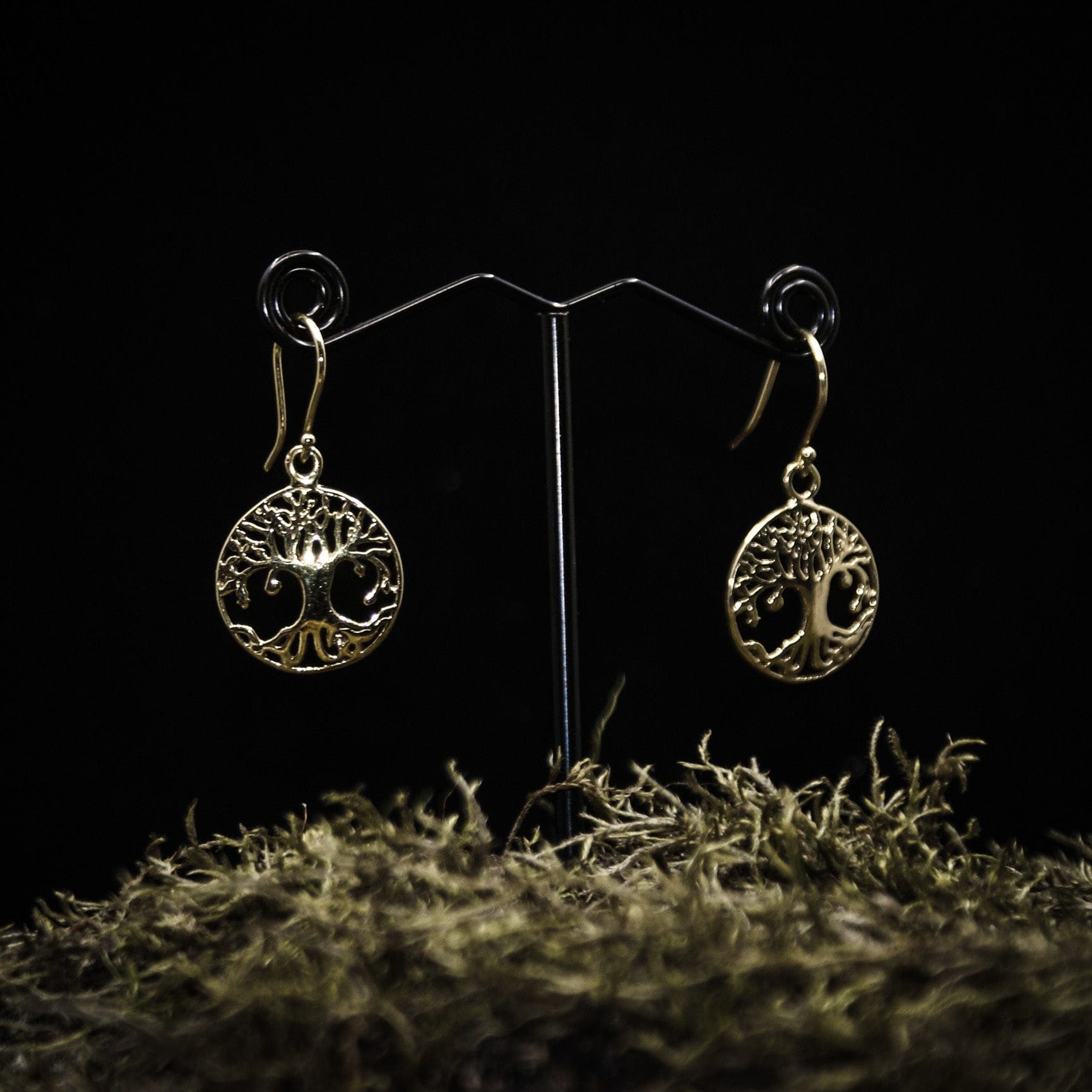 Ett par guldpläterade örhängen med Yggdrasilträd motiv, visade på en T-ställ mot mörkgrå bakgrund. Detaljerat trädsymbol med cirkulär ram och fligréprytninga i mässing.