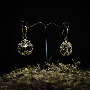 Ett par guldpläterade örhängen med Yggdrasilträd motiv, visade på en T-ställ mot mörkgrå bakgrund. Detaljerat trädsymbol med cirkulär ram och fligréprytninga i mässing.