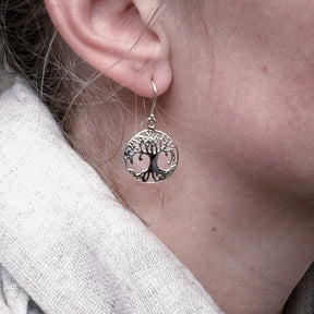 Närbild av en silverpläterad Yggdrasil örhänge som bärs av en kvinna. Visar hur örhänget hänger från örat med detaljer av träd motiv i cirkulär ram.