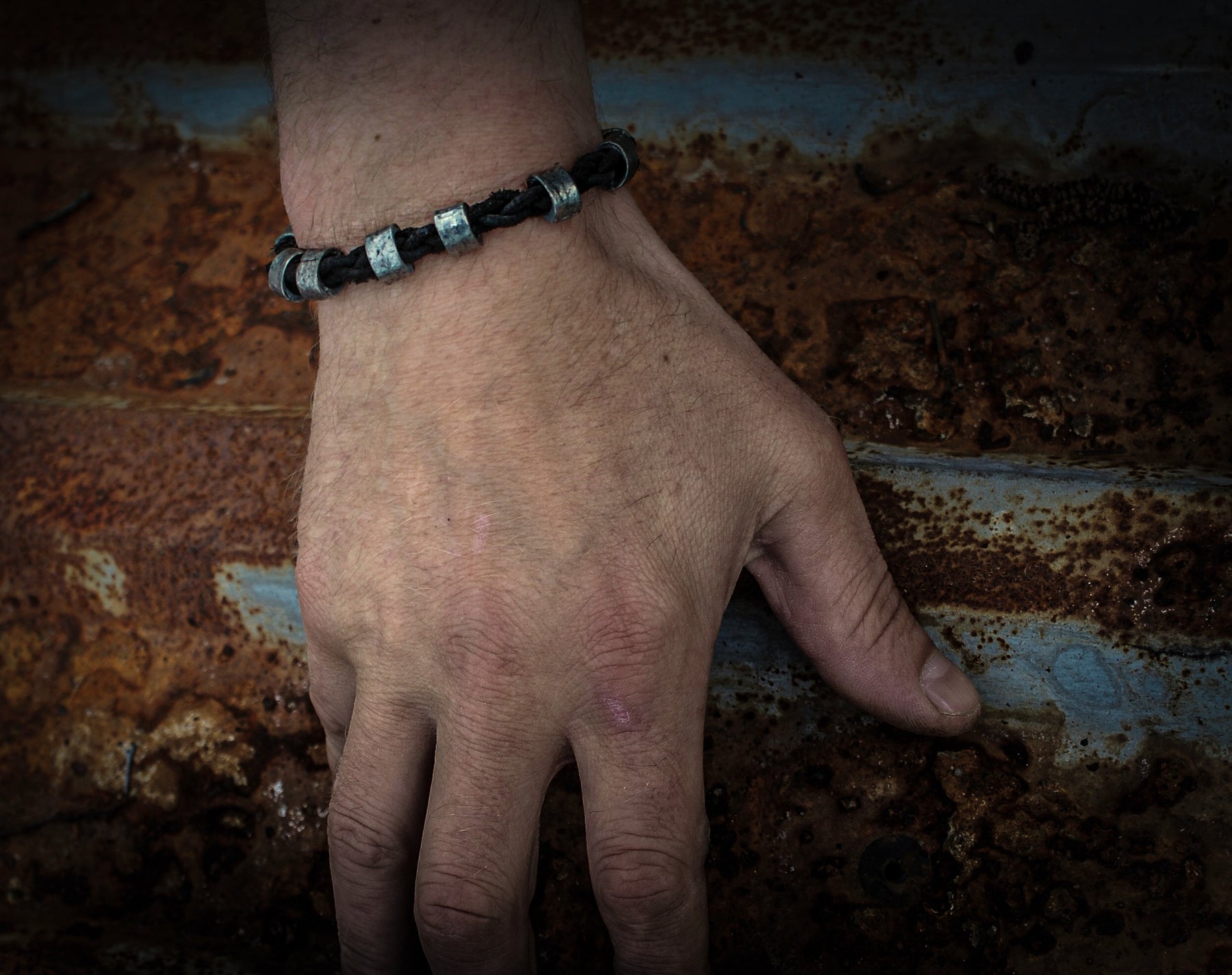 Vikingaarmbandet bärat på mans handled mot industriell rostmetall bakgrund med blå och rostiga patina, visar den flätade lädern och metallkulorna i daglig användning.