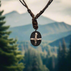 Halsband Byzans - enkelt bysantinskt korssmycke i tenn och naturfiber, upphängt i brunt läderband. Fotografi tagen mot en bergig skogslandskap bakgrund.