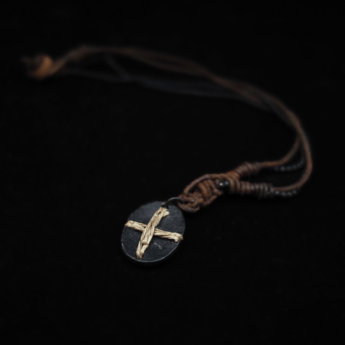 Närbild på Halsband Byzans-smycket, visar det dunkla ovalformade hänget med ljust korssmycke inlagt, upphängt i brunt läderband mot mörk bakgrund.