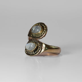Mässing spiralring med månstenar. Två blekt vita stenar med subtil blå-vit opalescens. Reptvinn-detaljer på mässingsfattningen visar traditionell vikinginspirerad hantverk. Ljus bakgrund framhäver stenernas glans.