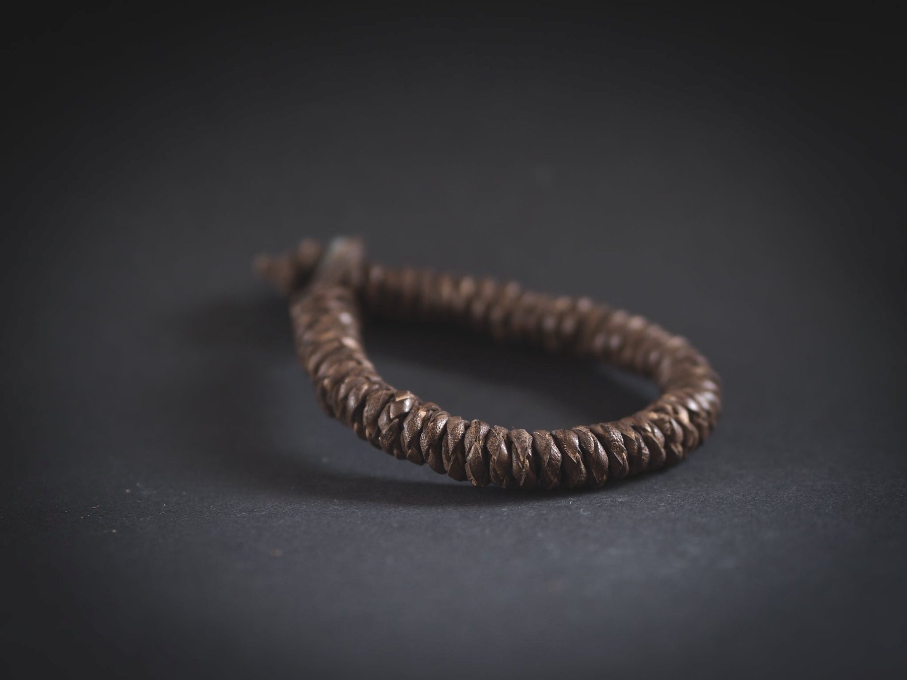 Produktfoto av brunt brokk-armband fotograferat mot mörkgrå bakgrund. Visar väl den täta flätningen och metallknappslutningen.