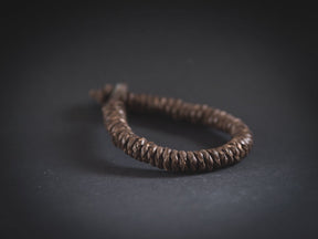 Produktfoto av brunt brokk-armband fotograferat mot mörkgrå bakgrund. Visar väl den täta flätningen och metallknappslutningen.