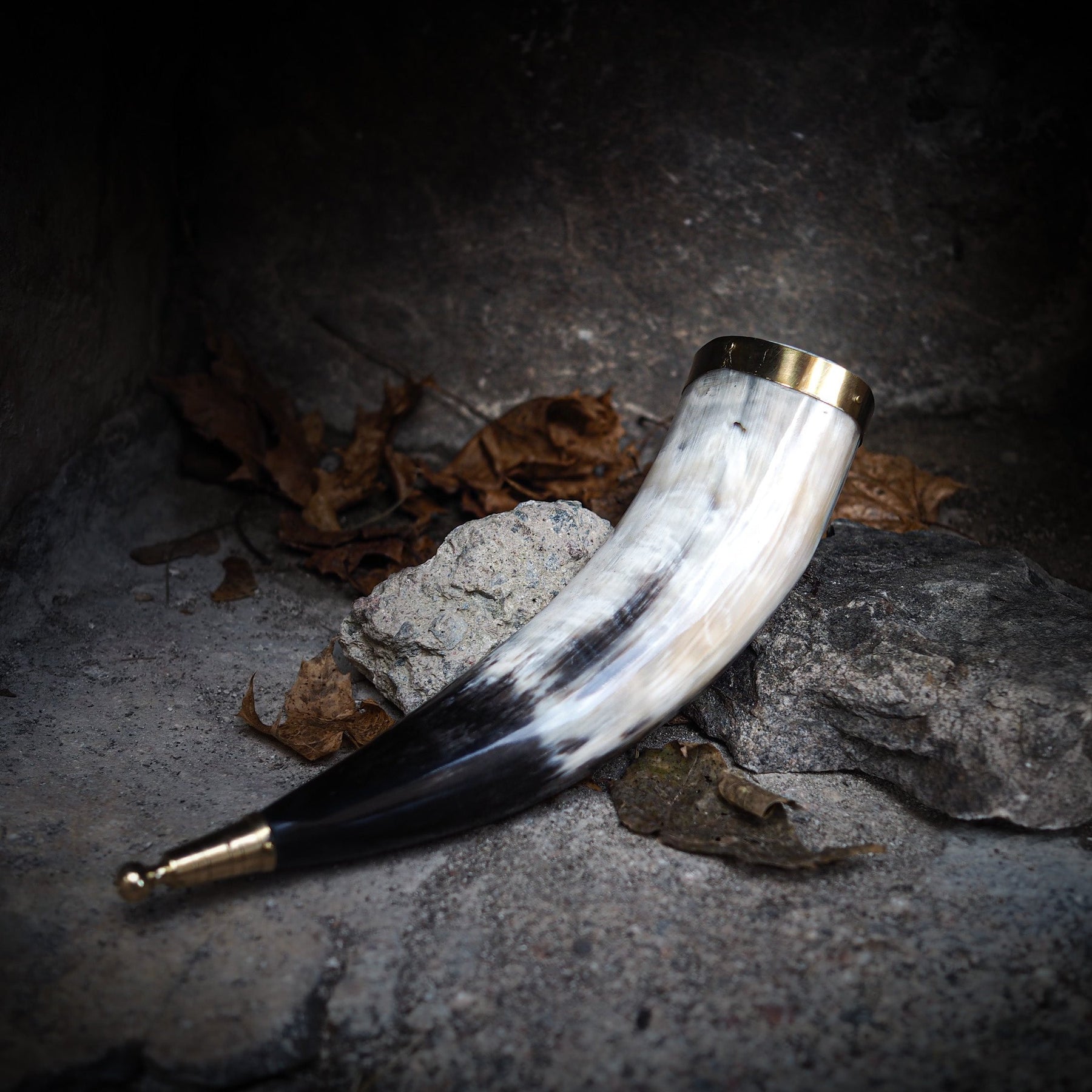 Ljust elfenbenskräm dryckeshorn av polerat horn med mässingsbeslag, stående på grå sten med höstlöv