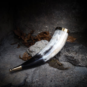 Ljust elfenbenskräm dryckeshorn av polerat horn med mässingsbeslag, stående på grå sten med höstlöv