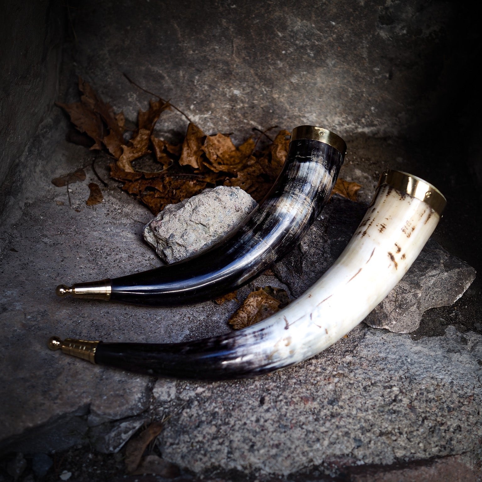 Tre dryckeshorn av polerat horn med mässingsdekor i olika färger från elfenbenskräm till mörkblå, visade på steingrund med höstlöv och natursten