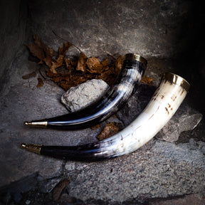 Tre dryckeshorn av polerat horn med mässingsdekor i olika färger från elfenbenskräm till mörkblå, visade på steingrund med höstlöv och natursten