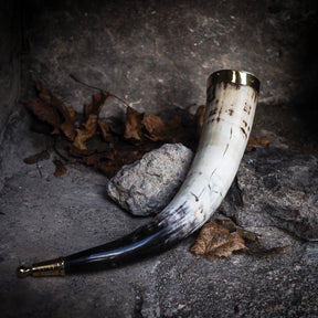 Dryckeshorn med snidad djurhuvudbase på naturlig bakgrund. Visar mässingsprydning, naturligt horn, och vikingarsnidad animalisk dekor med sten- och naturaccessoarer.