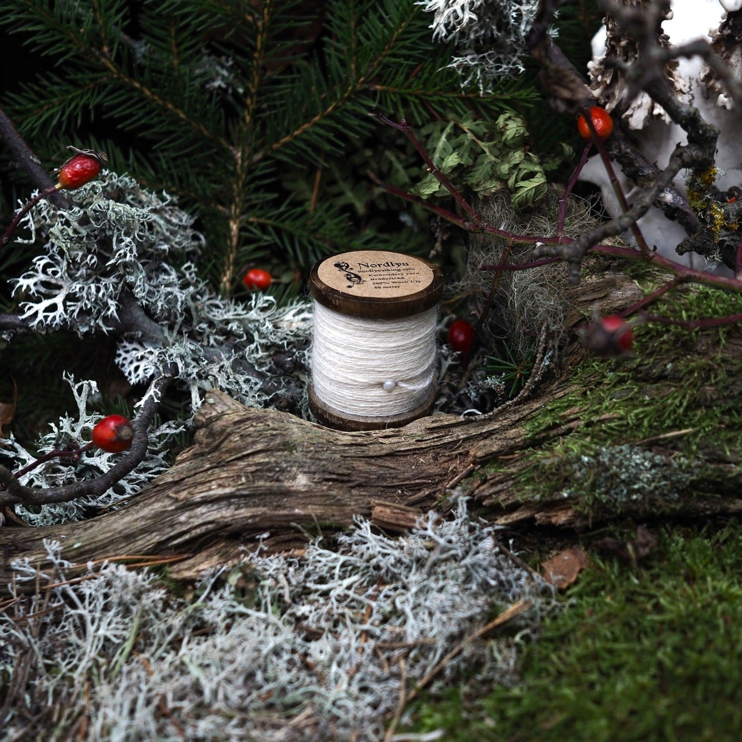 Naturvit brodyrtråd i 100% ull på träspole, placerad på naturlig mosslik miljö. Cirka 50 meter tråd för broderi och traditionellt handarbete. Fotograferad i utomhusmiljö.
